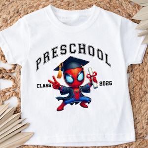 Puede incluir: Una camiseta blanca de manga corta con un gr&aacute;fico de un superh&eacute;roe de dibujos animados con birrete y toga de graduaci&oacute;n negros, sosteniendo un diploma. El texto dice "PRESCHOOL" en un arco arriba y "CLASS 2026" a ambos lados del personaje.