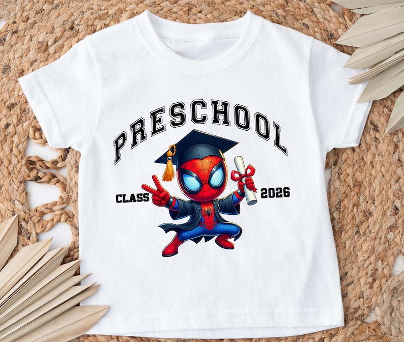 Puede incluir: Una camiseta blanca de manga corta con un gr&aacute;fico de un superh&eacute;roe de dibujos animados con birrete y toga de graduaci&oacute;n negros, sosteniendo un diploma. El texto dice "PRESCHOOL" en un arco arriba y "CLASS 2026" a ambos lados del personaje.