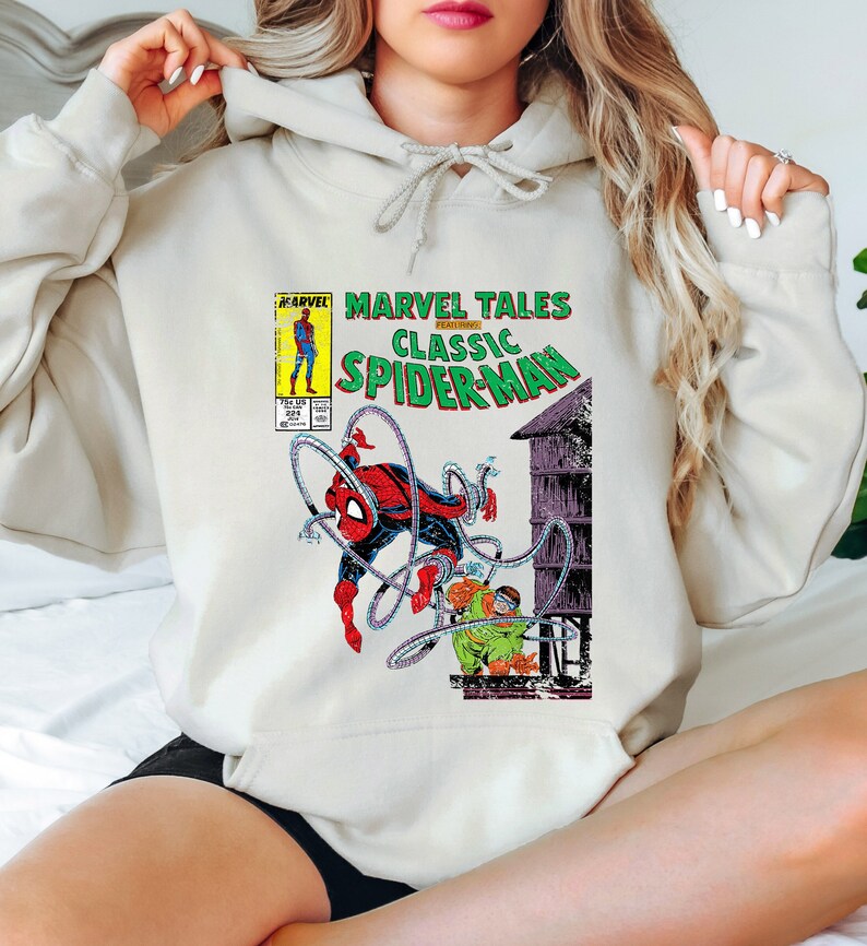 May include: Cream-colored hoodie featuring a Marvel Tales Classic Spider-Man comic book cover design. The graphic includes Spider-Man in action, with text in green, red, and yellow. The hoodie has a drawstring hood.