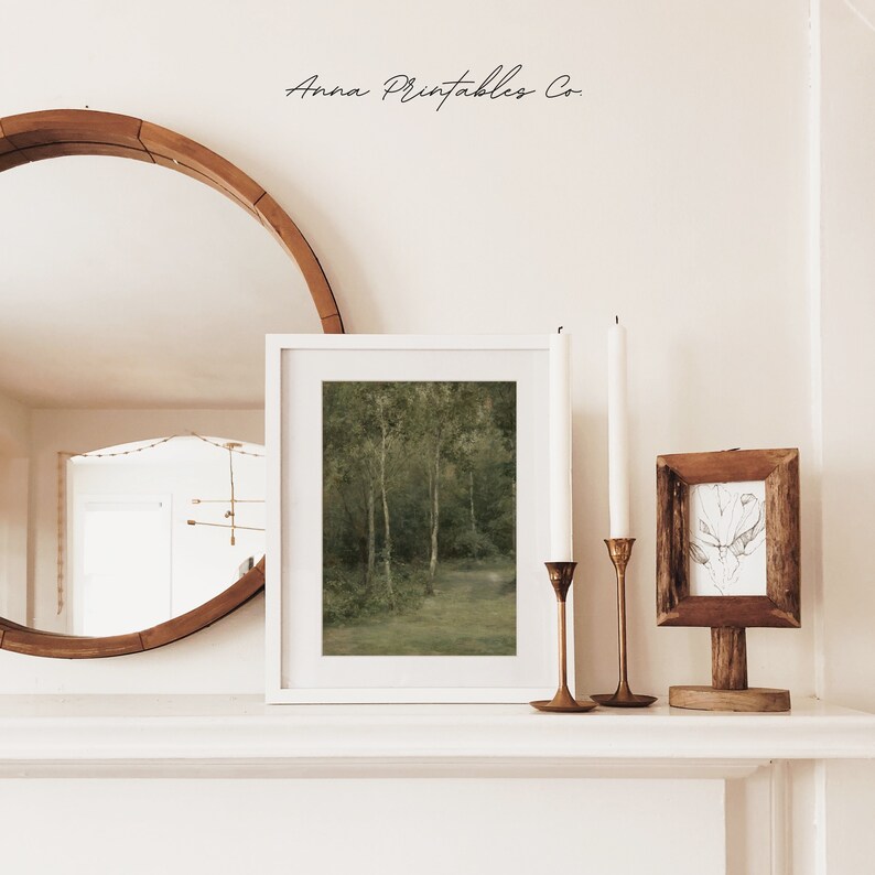 May include: A white framed print of a green forest scene with trees and a path. The print is on a white shelf with two candlesticks and a small wooden frame with a drawing.