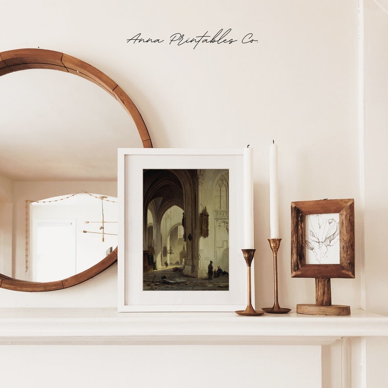 May include: A white framed print of a painting depicting the interior of a church with arched ceilings and stone walls. The painting is displayed on a white shelf with two candlesticks and a small wooden frame with a drawing.