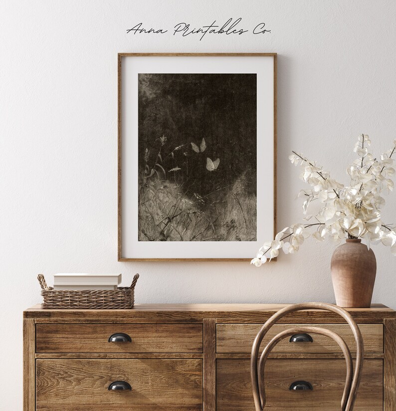 May include: A sepia-toned framed print featuring a dark, textured background with three butterflies and delicate plant life. The print is displayed above a wooden dresser with a woven basket and a vase of white flowers.