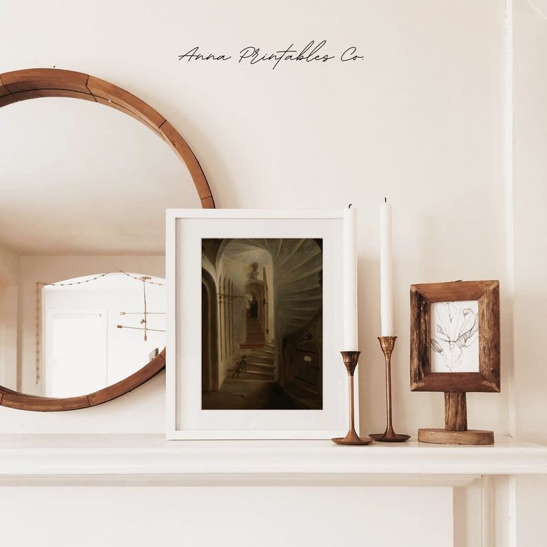 May include: A white framed print of a painting depicting a dark, winding staircase with a person walking up the stairs. The print is displayed on a white shelf with two candlesticks and a small wooden frame with a drawing inside.