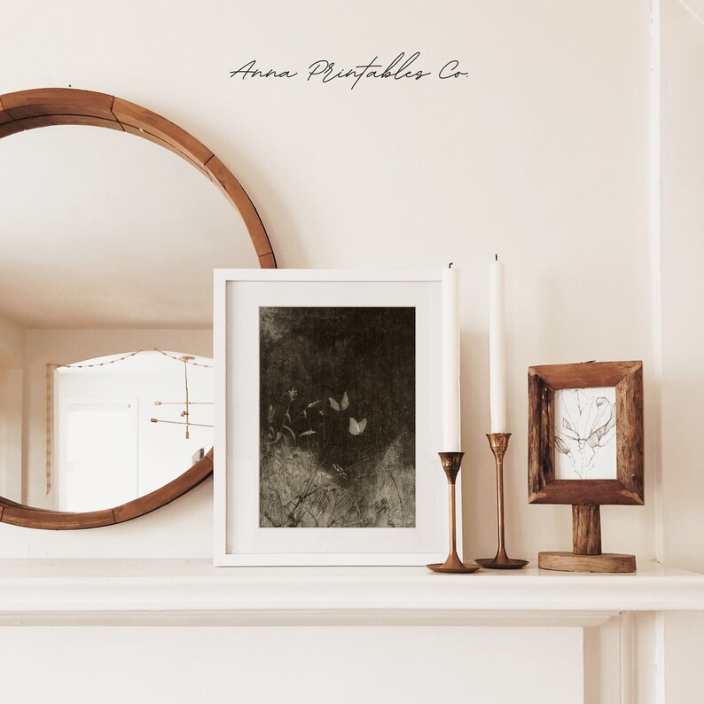 May include: A white framed print with a dark, moody botanical illustration featuring butterflies and plants. Two brass candle holders with white candles and a small framed sketch are also on display. The text "Anna Printables Co." is at the top.