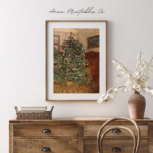 Pode incluir: Uma pintura vintage de uma &aacute;rvore de Natal decorada com ornamentos e luzes em uma sala com um arm&aacute;rio de madeira e um tapete no ch&atilde;o. A pintura &eacute; emoldurada em uma moldura de madeira clara.