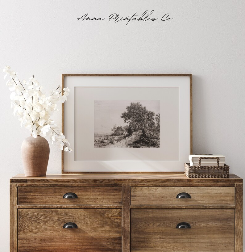 May include: A black and white photograph of a landscape with a large tree in the foreground. The photograph is framed in a wooden frame and is displayed on a wooden dresser with two drawers.