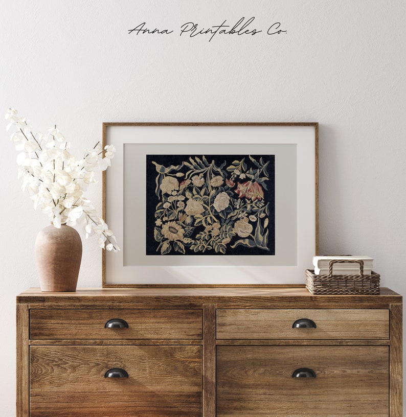 May include: A framed print of a vintage floral tapestry with a dark blue background and white, yellow, and red flowers. The print is in a wooden frame and is hanging on a white wall.