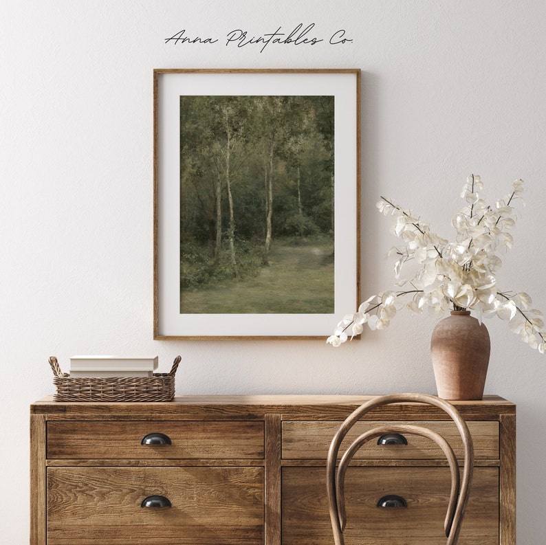 May include: A framed print of a painting depicting a forest scene with tall trees and a path leading into the woods. The print is in a light wood frame and is hanging on a white wall above a wooden dresser.