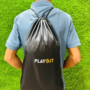 Person viewed from behind wearing a black drawstring backpack with the “PLAYBIT” logo in white and yellow, standing on bright green artificial grass.