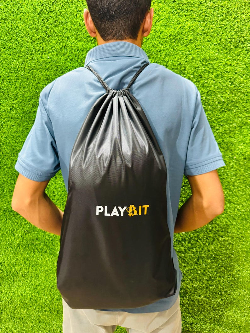 Person viewed from behind wearing a black drawstring backpack with the “PLAYBIT” logo in white and yellow, standing on bright green artificial grass.