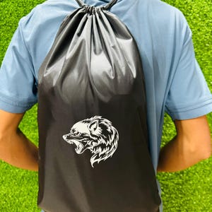 Person wearing a black drawstring backpack with a white wolf head graphic, photographed from behind while standing on bright green artificial grass.