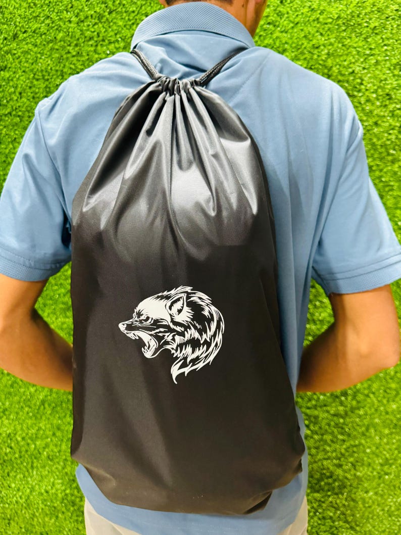 Person wearing a black drawstring backpack with a white wolf head graphic, photographed from behind while standing on bright green artificial grass.
