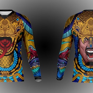 May include: A long-sleeved shirt with a colorful design featuring a golden snake head with a red tongue and a warrior's face with a blue and gold background. The shirt has the text "VIIA" on the bottom.
