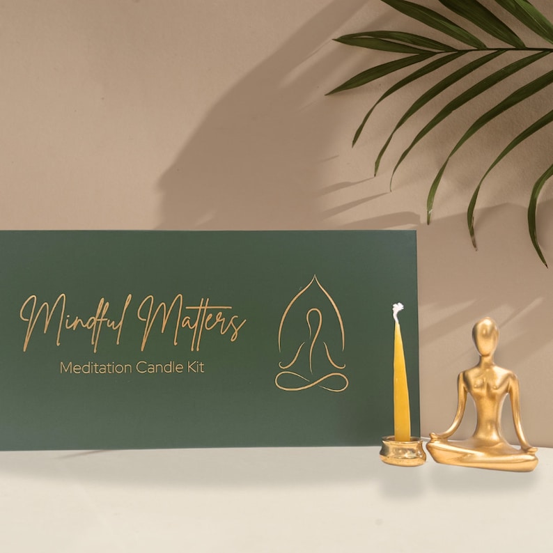 May include: A green box with gold lettering that reads "Mindful Matters Meditation Candle Kit". A gold outline of a person sitting in a meditative pose is on the right side of the box. A small gold statue of a person sitting in a meditative pose and a gold candle holder with a yellow candle are in front of the box.