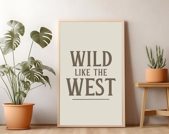 May include: A beige framed print with the text "WILD LIKE THE WEST" in brown lettering. The print is hanging on a white wall with a green plant in a terracotta pot in the foreground.