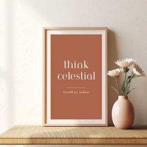 Könnte beinhalten: Ein gerahmter Druck mit terracottafarbenem Hintergrund und weißem Text, der "think celestial" lautet, mit dem Namen "russell m. nelson" darunter. Der Druck liegt auf einer Holzoberfläche mit einer Vase mit weißen Blumen im Hintergrund.