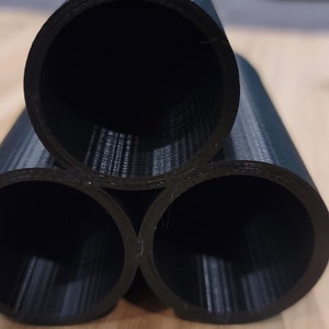 May include: Three black plastic tubes stacked in a triangular shape on a wooden surface. The tubes are hollow and have a smooth, slightly textured exterior.