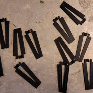 May include: Ten black plastic pieces with a V-shaped opening in the center. The pieces are arranged in a scattered pattern on a gray surface.