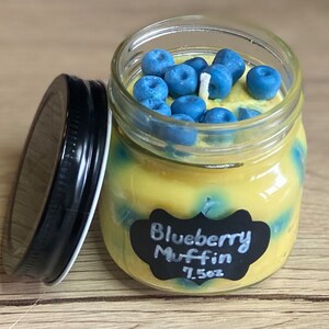 May include: A glass jar candle with a black label that reads "Blueberry Muffin 7.5oz". The candle is topped with blue blueberry-shaped wax and a white wick.