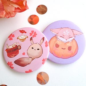 Puede incluir: Dos botones redondos de color rosa y morado con ilustraciones de animales de dibujos animados. El botón rosa presenta un conejo marrón con un sombrero rojo y blanco y el botón morado presenta un zorro rosa en una calabaza.