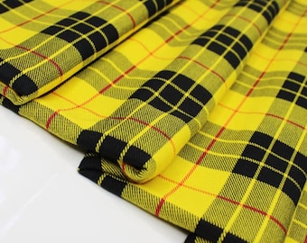 Op de afbeelding: Close-up van gele geruite stof met zwarte en rode accenten. De stof is opgevouwen en toont de herhalende vierkanten en lijnen van het patroon. De heldere gele kleur domineert en creëert een visueel opvallend textiel.