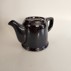 May include: A dark brown ceramic teapot with a shiny glaze. The teapot has a rounded body and a spout on the side. The handle is on the opposite side of the spout.