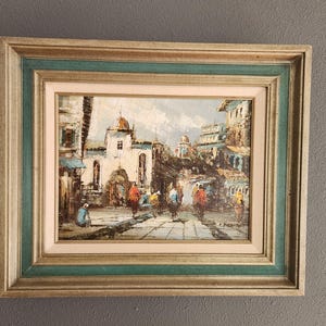 May include: A framed oil painting depicting a street scene in a European city. The painting features a white building with a red roof, and people walking on a cobblestone street. The painting is framed in a wooden frame with a gold trim.