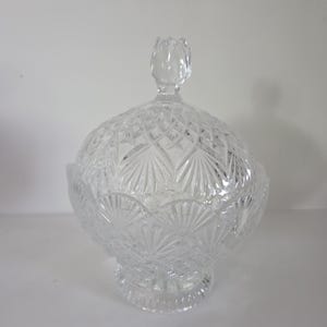 May include: A clear, cut-glass candy dish with a lid. The dish has a round, pedestal base and a domed lid with a flower-shaped handle. The glass is intricately cut with a diamond and fan pattern, creating a sparkling effect.