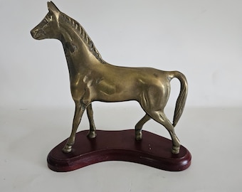 May include: A brass horse figurine standing on a wooden base. The horse is in a walking pose with its head turned to the side. The base is a dark brown color and has a slightly curved shape.