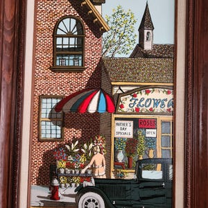 May include: A framed painting depicts a flower shop scene. A woman arranges flowers in a black truck. The shop has a red, white, and blue striped awning. The sign reads "FLOWERS" and "MOTHER'S DAY SPECIALS". A red fire hydrant is visible.