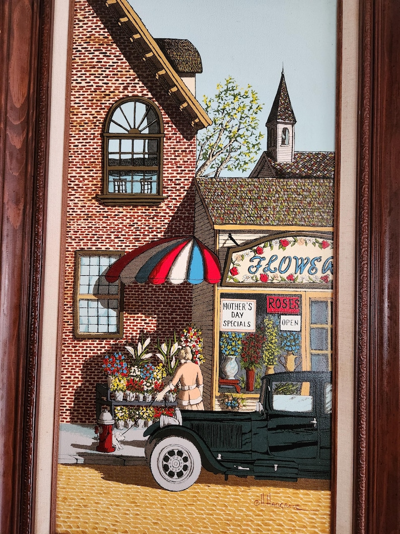 May include: A framed painting depicts a flower shop scene. A woman arranges flowers in a black truck. The shop has a red, white, and blue striped awning. The sign reads "FLOWERS" and "MOTHER'S DAY SPECIALS". A red fire hydrant is visible.