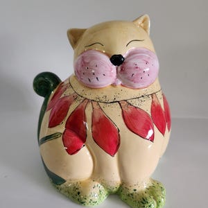 May include: A ceramic cat figurine with a cream-colored body, pink cheeks, and a black nose. The cat has red and pink floral accents around its neck and green accents on its feet and tail. The cat is sitting.