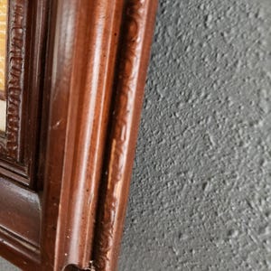 May include: Close-up of a dark brown wooden picture frame with ornate detailing. The frame has a glossy finish and is set against a textured gray wall. The corner of the frame shows slight wear.