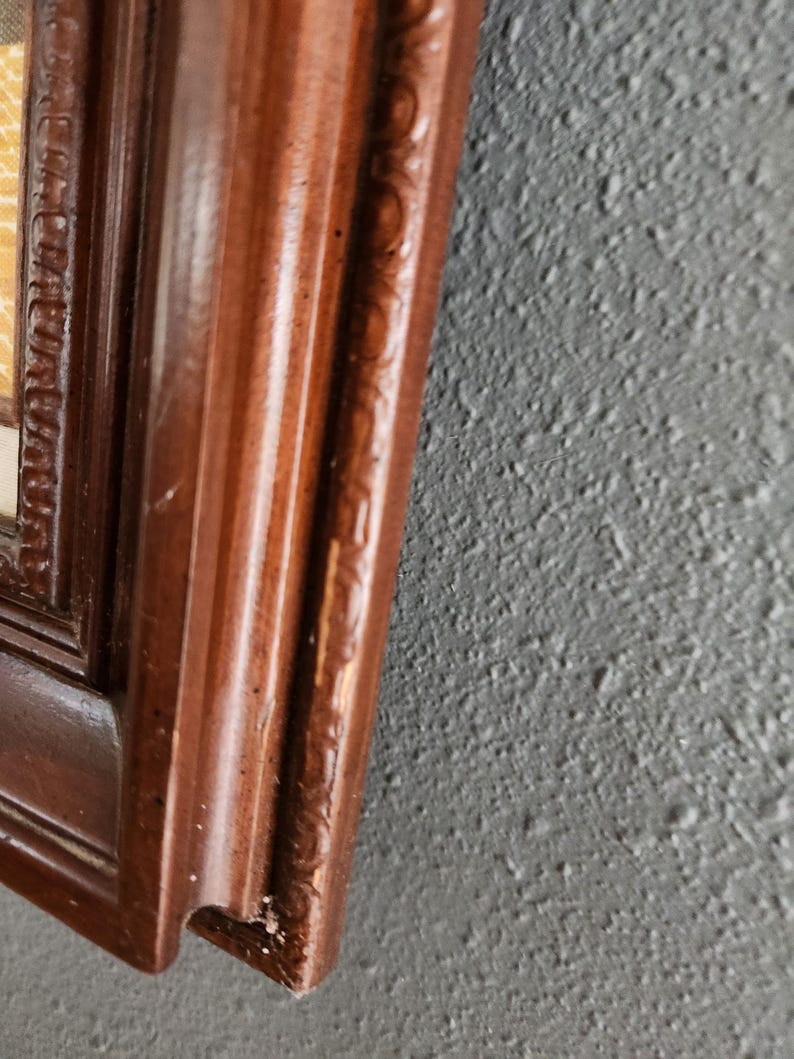 May include: Close-up of a dark brown wooden picture frame with ornate detailing. The frame has a glossy finish and is set against a textured gray wall. The corner of the frame shows slight wear.
