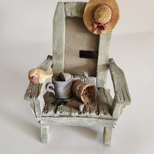 May include: A decorative miniature Adirondack chair picture frame with a weathered, rustic appearance. The frame features a straw hat with a pink bow, a small bird, a watering can, a flower pot with soil, and gardening tools. The frame is approximately 6 inches tall.