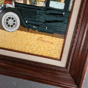 May include: An artistic framed print depicting a vintage car parked in front of a flower shop. The car is dark green with white tires, and the shop features colorful flowers. The frame is dark brown.