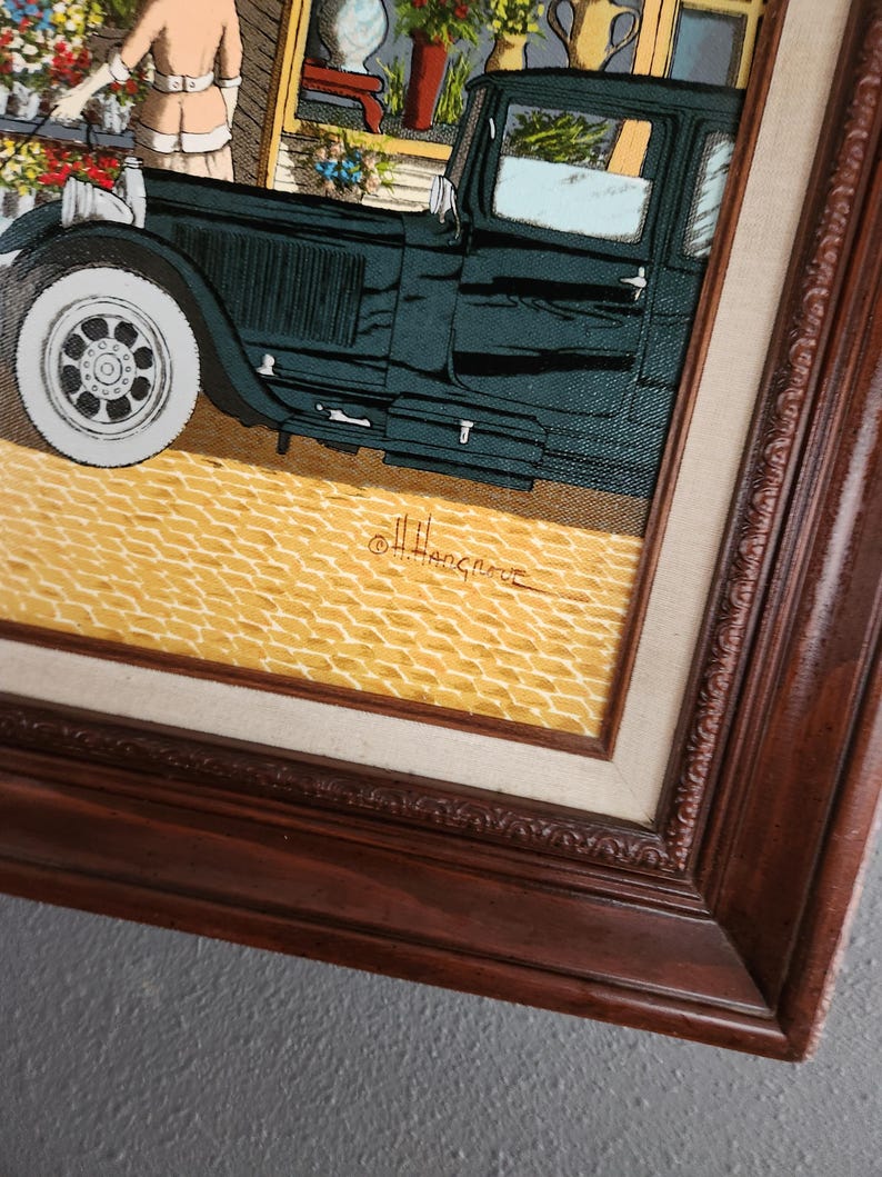 May include: An artistic framed print depicting a vintage car parked in front of a flower shop. The car is dark green with white tires, and the shop features colorful flowers. The frame is dark brown.