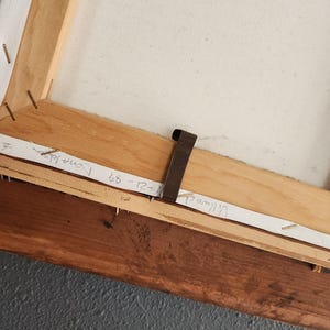 May include: The back of a stretched canvas artwork, showing the wooden frame and canvas. The frame is made of light brown wood, with staples securing the canvas. Handwritten text on paper strips includes names and dates.