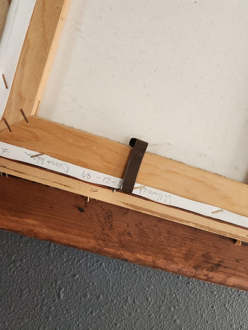 May include: The back of a stretched canvas artwork, showing the wooden frame and canvas. The frame is made of light brown wood, with staples securing the canvas. Handwritten text on paper strips includes names and dates.