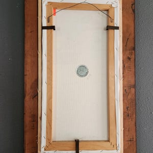 May include: Back of a framed painting with a wooden frame. The canvas is stretched and stapled to a wooden frame. The back of the canvas has a metal wire for hanging. The frame is a dark brown color.