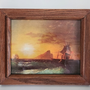 May include: A framed painting of a seascape with two ships sailing towards a rocky island at sunset. The sky is a vibrant orange and yellow, with clouds in the distance. The water is a deep blue, with whitecaps on the waves.