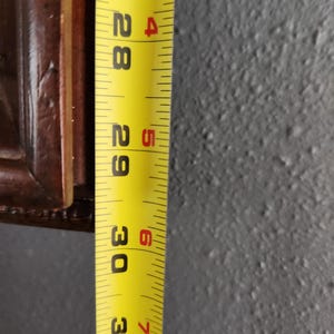 May include: A yellow measuring tape showing the 28 to 31-inch marks. The tape is next to a dark brown wooden frame. The background is a textured gray wall.