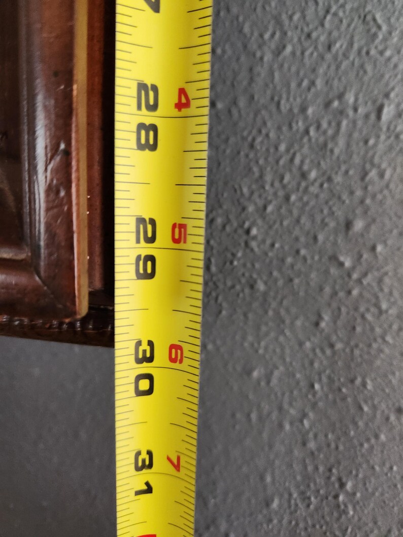 May include: A yellow measuring tape showing the 28 to 31-inch marks. The tape is next to a dark brown wooden frame. The background is a textured gray wall.