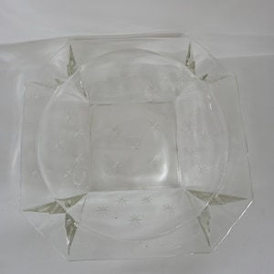 May include: Clear glass ashtray with a square shape and a circular interior. The ashtray features a starburst pattern etched into the glass. The corners of the ashtray have pointed, triangular extensions.