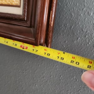 May include: A framed artwork with a dark brown wooden frame. A yellow measuring tape shows the frame is approximately 18 inches long. The artwork inside the frame has a light beige textured surface.
