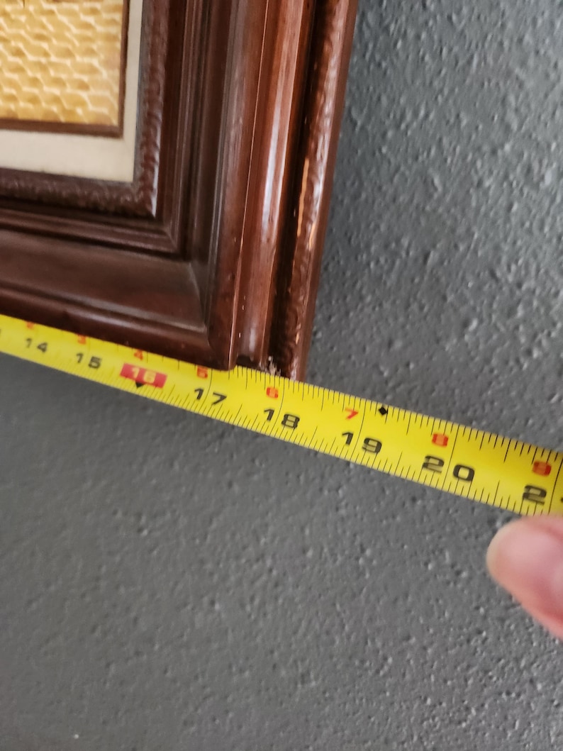May include: A framed artwork with a dark brown wooden frame. A yellow measuring tape shows the frame is approximately 18 inches long. The artwork inside the frame has a light beige textured surface.