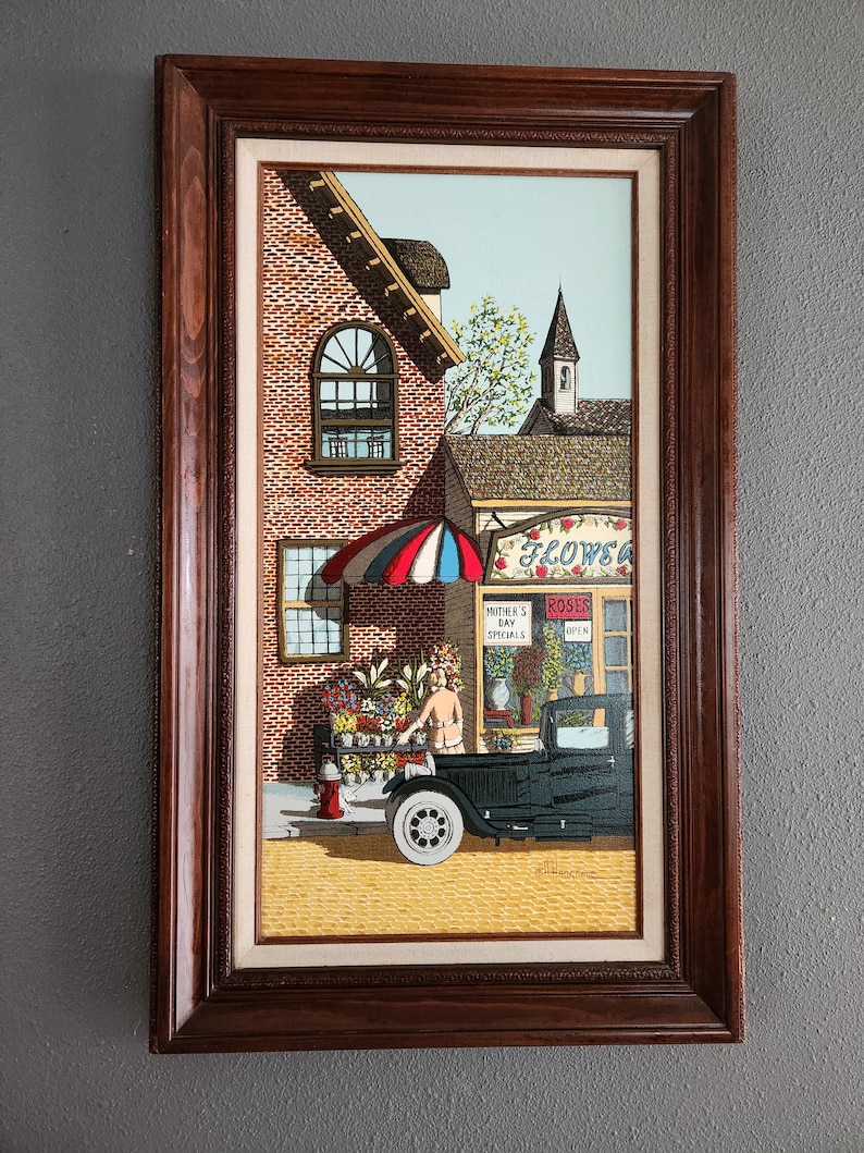 May include: Framed painting depicting a flower shop scene. The artwork features a brick building with a flower shop, a black vintage car, and a woman arranging flowers. The shop has a striped umbrella and the text "Mother's Day Specials".