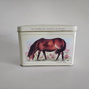 May include: A rectangular tin box with a cream-colored background. The box features a painting of a brown horse grazing in a field of pink flowers. The text "The Countryside Collection by Bristows" is printed on the lid.