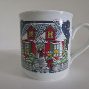 May include: A white ceramic mug featuring a colorful illustration of a red house decorated for the holidays. The scene includes a wreath on the door, a lamppost with a red bow, snow-covered roofs, and two decorated Christmas trees under a dark blue sky.