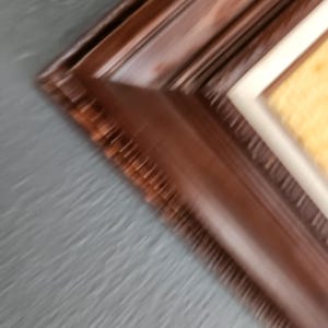 May include: Close-up of a dark brown wooden picture frame with a textured surface. The frame has a beveled edge and a cream-colored matting around the artwork. The background is a blurred gray.